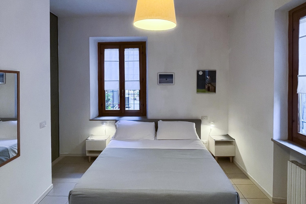 A bedroom with a large bed, two bedside tables, a hanging lamp, a window with a view, and a door.