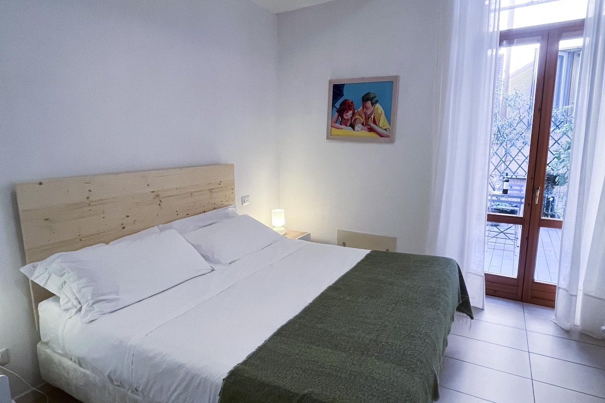 A bedroom with a wooden headboard, a bed with white linens, a green throw blanket, a nightstand with a lamp, and a painting on the wall.