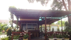 A covered outdoor seating area with a wooden structure, potted plants, and a green metal floor. - Coconut Home Resort (Bang Khonthi)