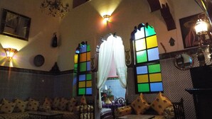 A room with a patterned wall, a sofa with patterned cushions, a chandelier, and a window with colorful panes. - Maison Adam (Fes)