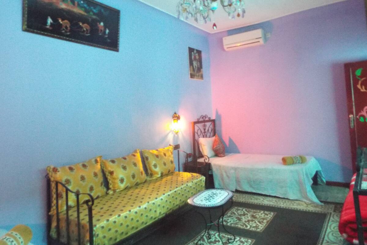 A room with a yellow sofa, a small table, a bed with a white bedspread, a chandelier, and a painting on the wall.