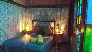 A room with a patterned wall, a bed with a blue bedspread, two pillows, a nightstand with a lamp, and a decorative lantern. - Maison Adam (Fes)