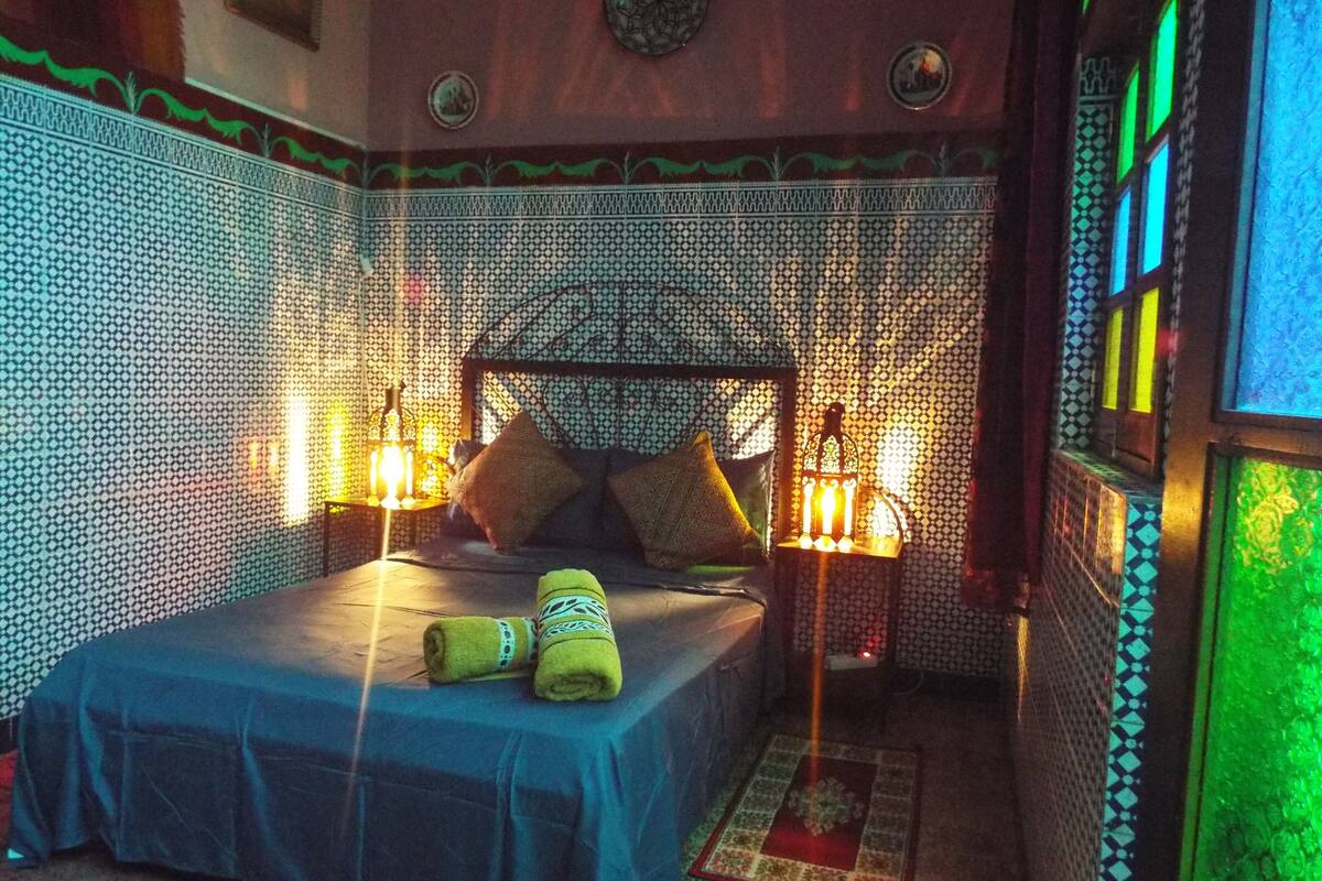 A room with a patterned wall, a bed with a blue bedspread, two pillows, a nightstand with a lamp, and a decorative lantern.