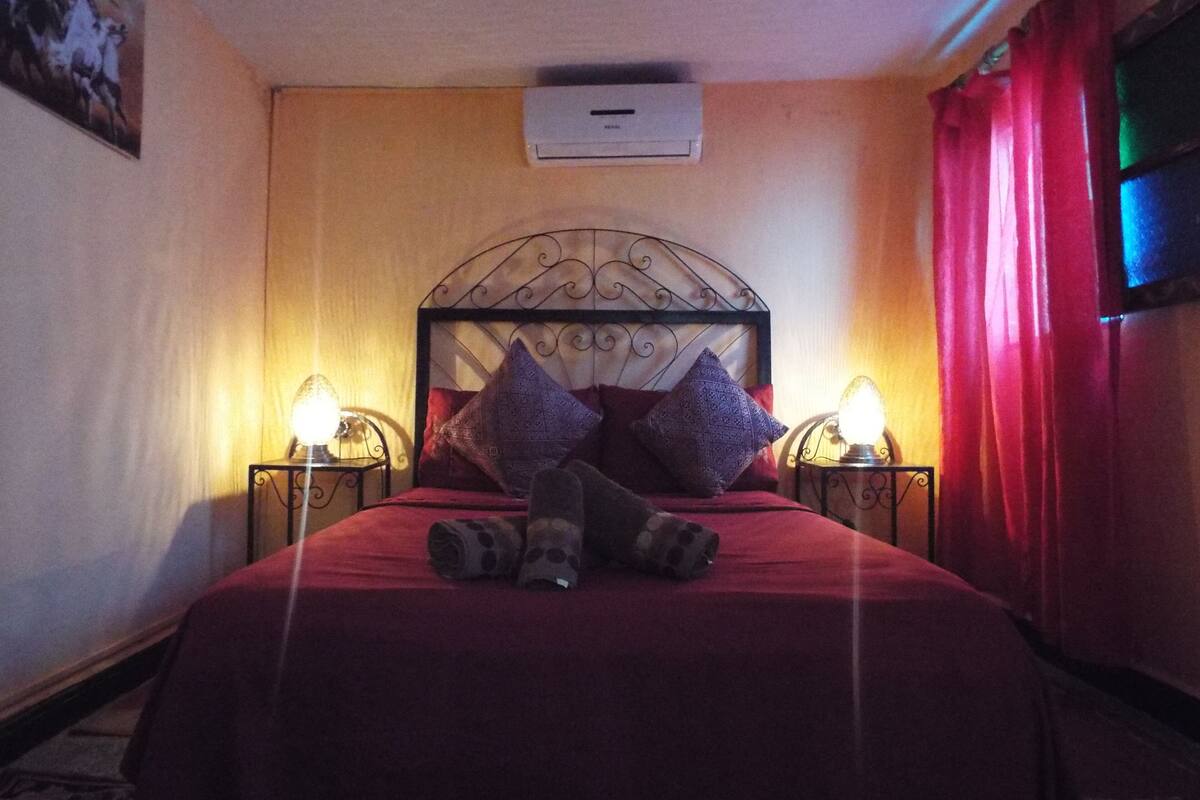 A bedroom with a bed, two bedside lamps, a wall-mounted air conditioner, and a red curtain.