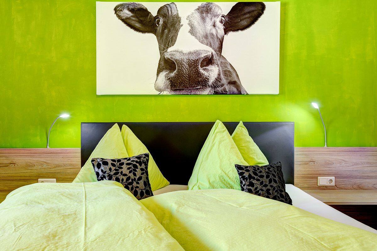 A bed with a green headboard, multiple pillows, and a wall-mounted artwork of a cow's face.