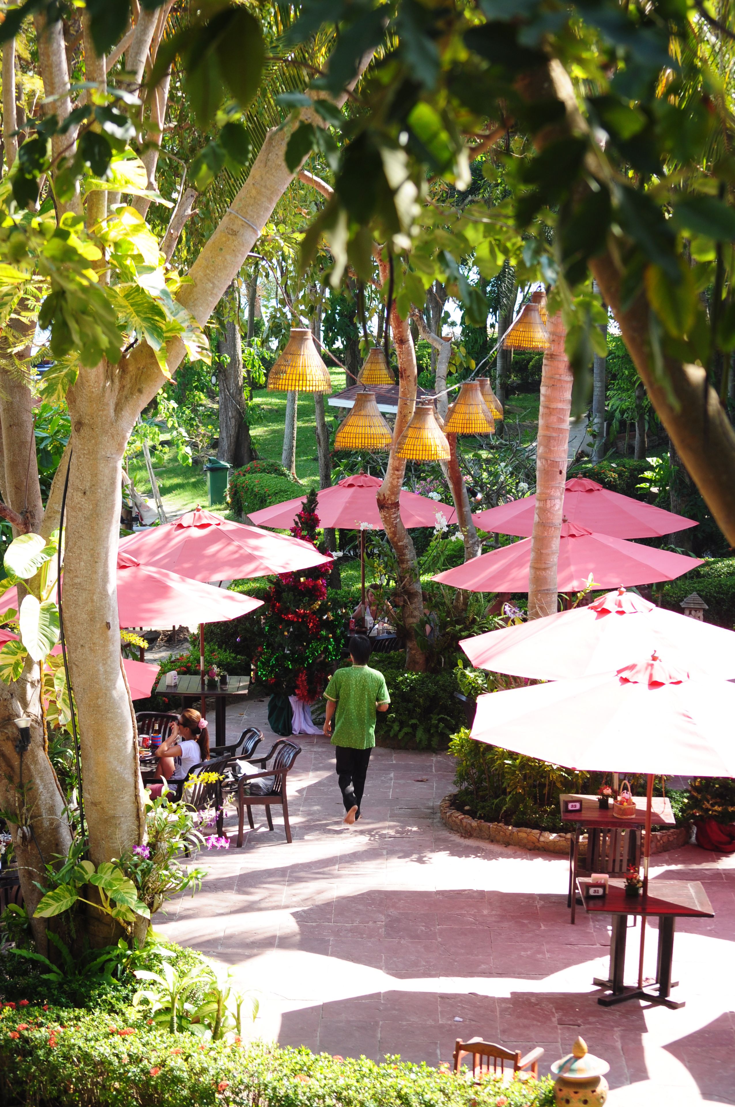 An outdoor patio area with tables and chairs under red umbrellas, surrounded by lush greenery and trees.