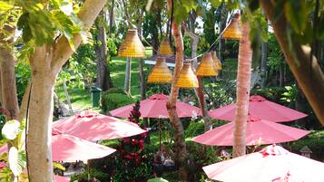 An outdoor patio area with tables and chairs under red umbrellas, surrounded by lush greenery and trees.
