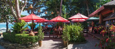 Un espace extérieur aménagé dans un style tropical, avec un sol en pierre, entouré d’une végétation luxuriante et agrémenté de parasols rouges.