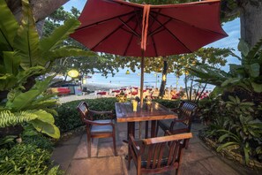 Un espace extérieur aménagé avec une table et des chaises, protégé par un parasol rouge, et offrant une vue sur une plage parsemée de parasols et de chaises.