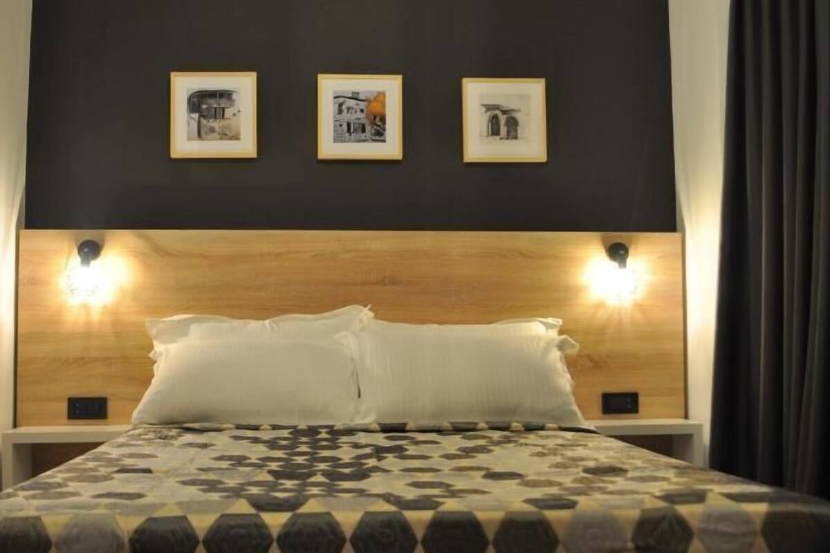 A hotel room with a wooden headboard, two wall-mounted lights, a bed with a patterned bedspread, and three framed pictures on the wall.