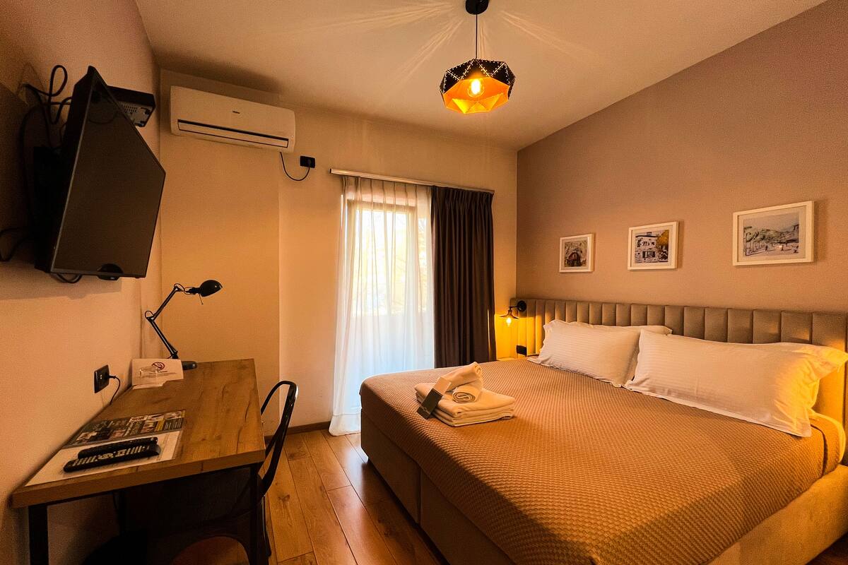 A hotel room with a bed, a desk with a computer, a TV, and a window with curtains.