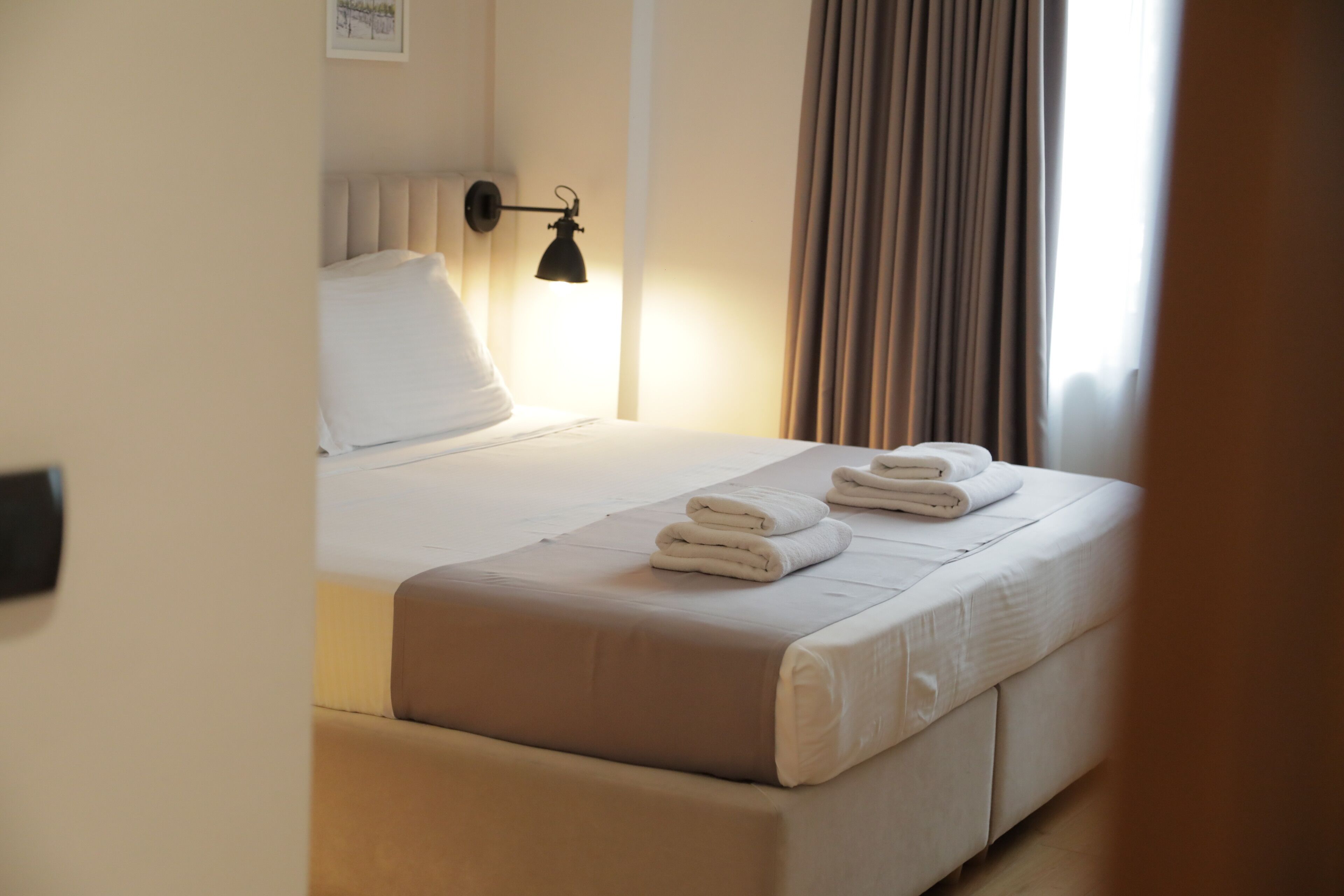 A neatly made bed with white linens and folded towels, a bedside lamp, and a window with curtains.