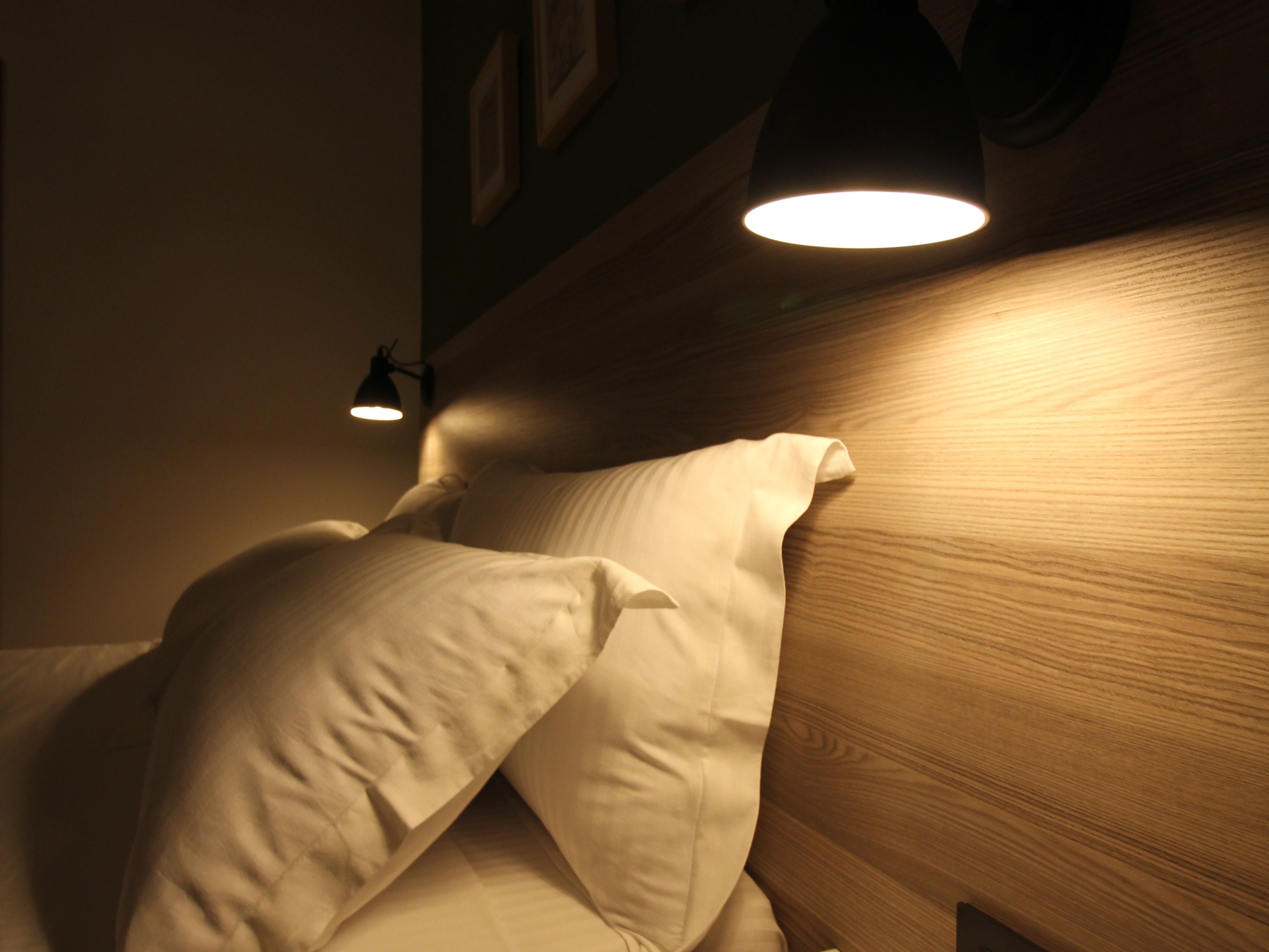 A bed with white linens and a wooden headboard. There is a wall-mounted light fixture above the bed.