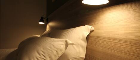 A bed with white linens and a wooden headboard. There is a wall-mounted light fixture above the bed.
