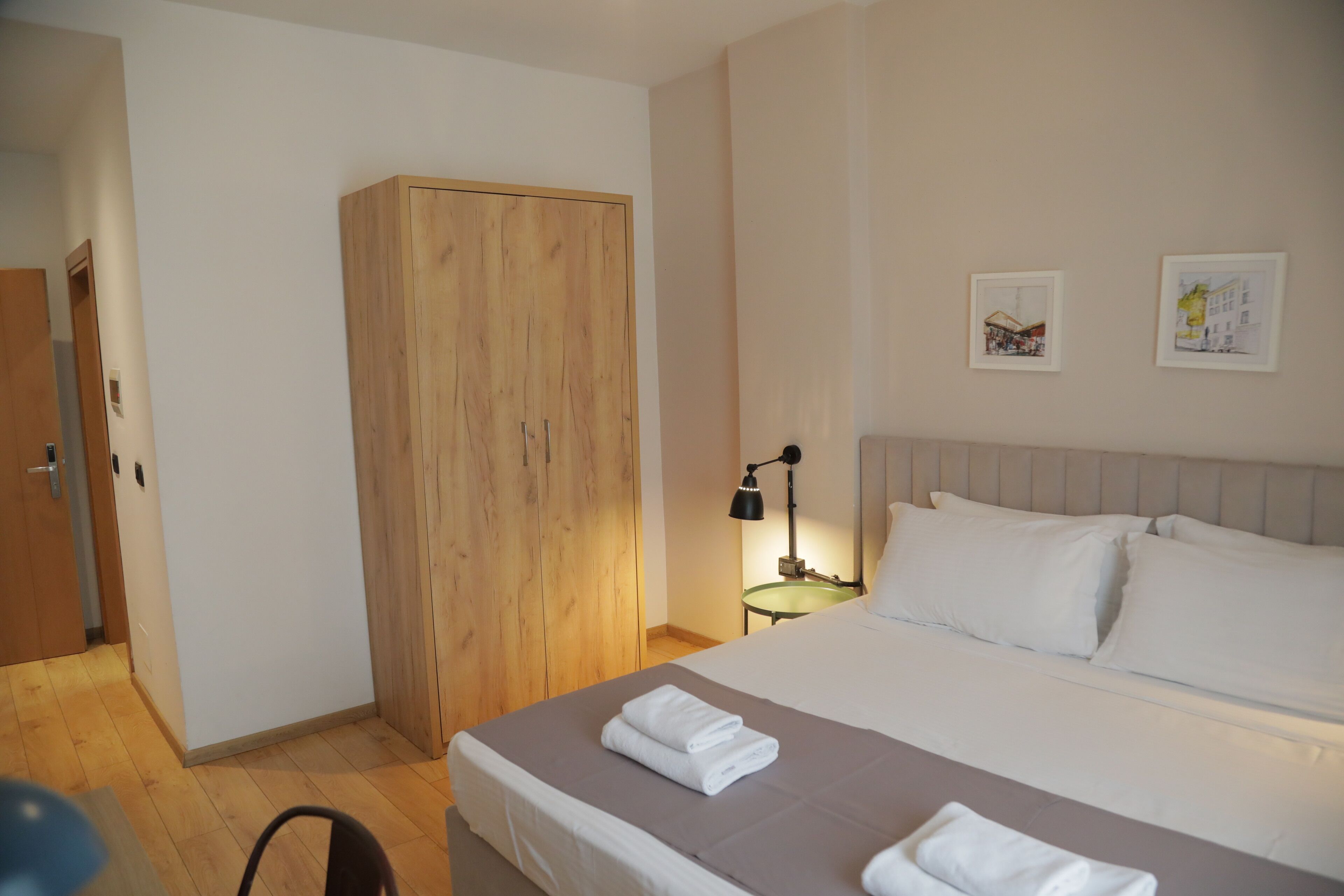 A hotel room with a bed, a wardrobe, a nightstand with a lamp, and two wall-mounted pictures.