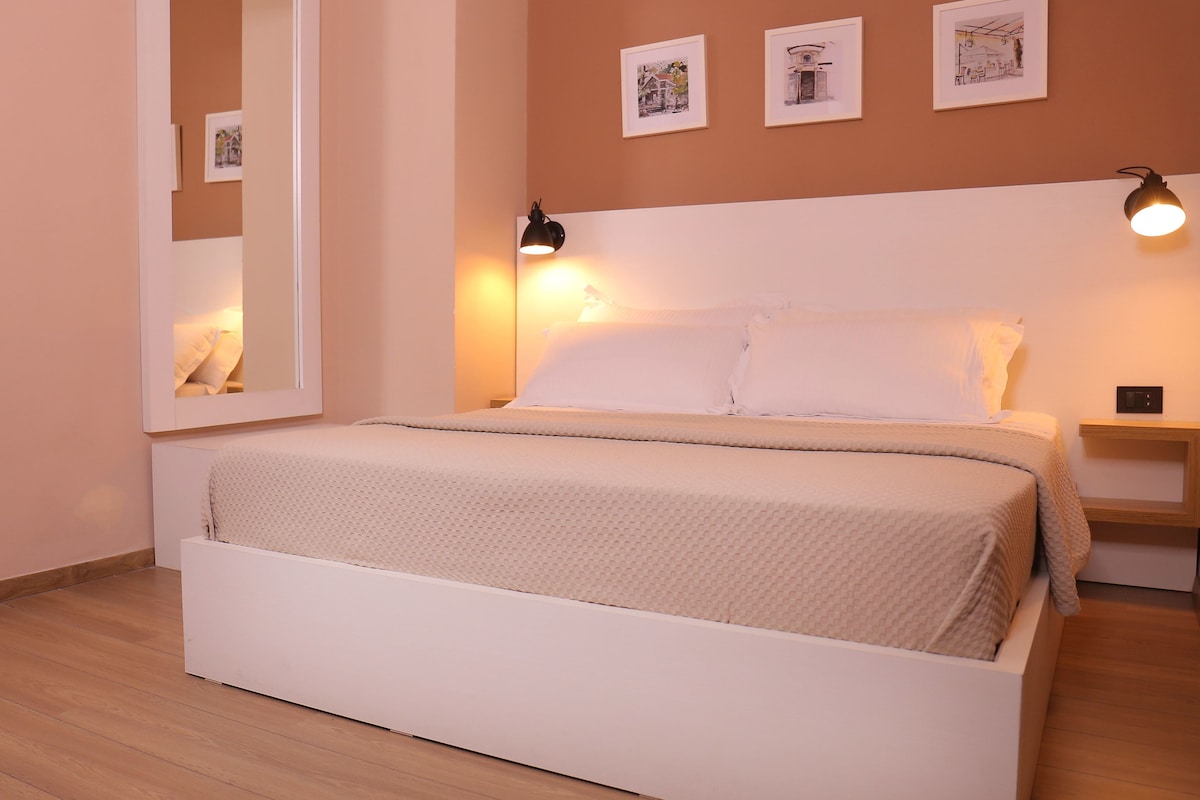 A bedroom with a bed, a bedside table, a nightstand, a wall-mounted lamp, and framed pictures on the wall.