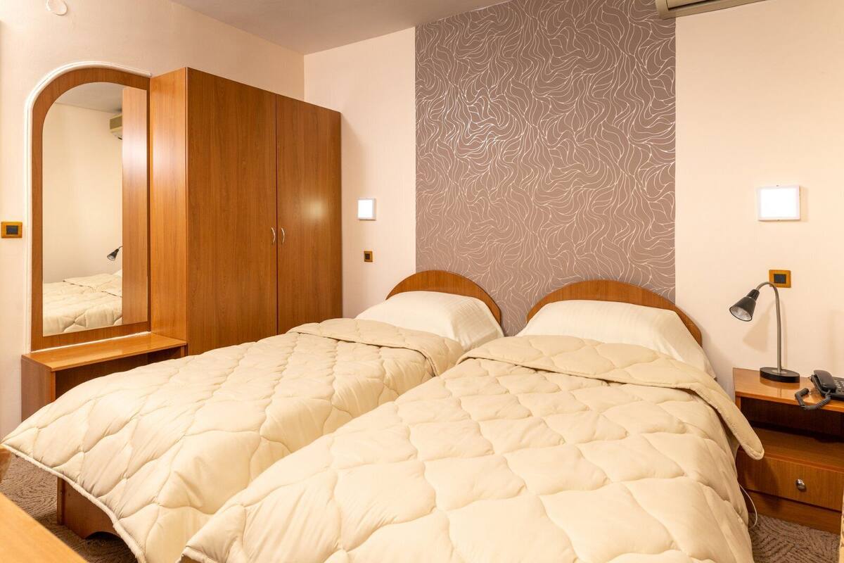 A double bed with a quilted duvet, a wooden wardrobe, a full-length mirror, a bedside table with a lamp, and a patterned wall.
