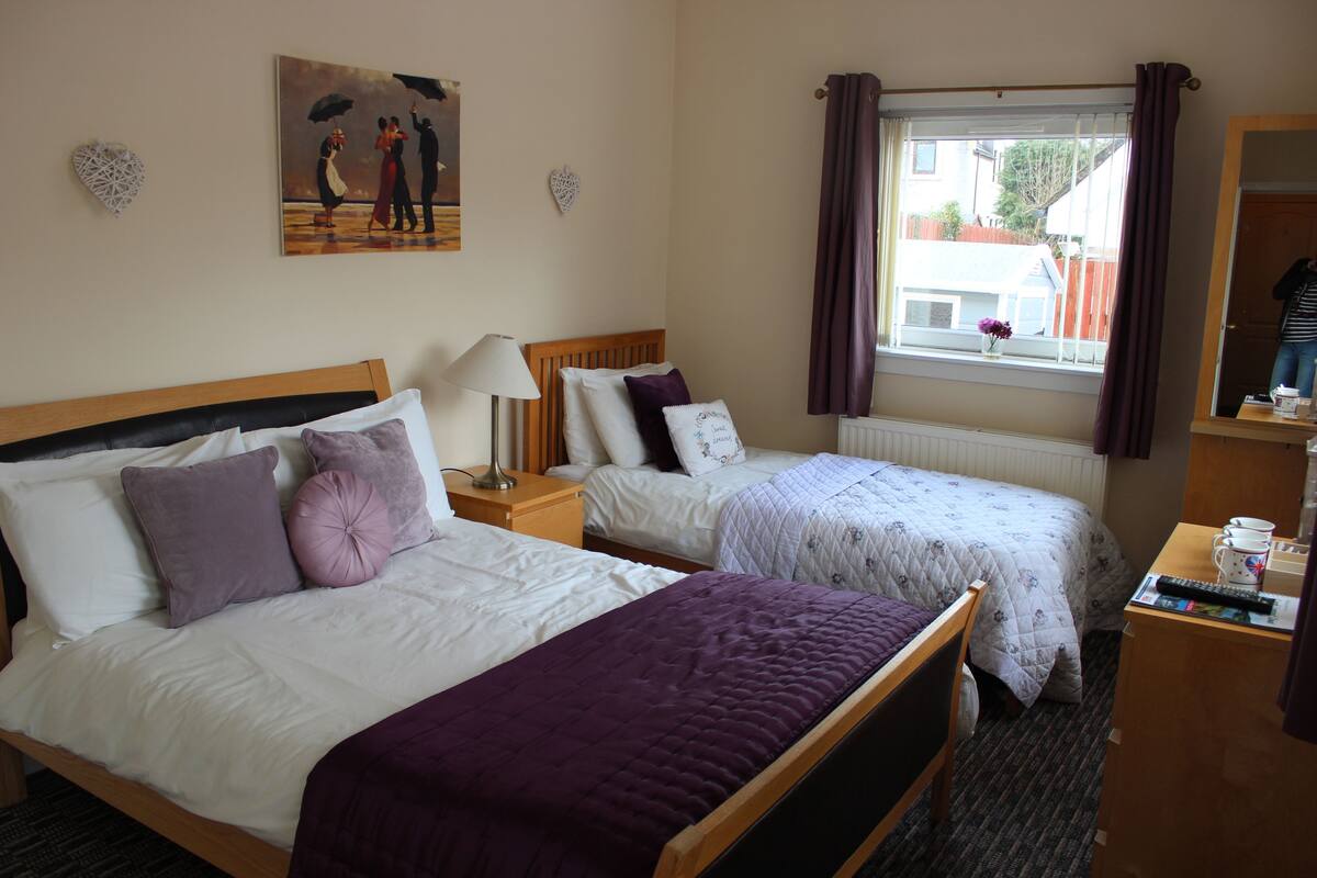 A hotel room with two beds, a nightstand with a lamp, a window with curtains, and a painting on the wall.