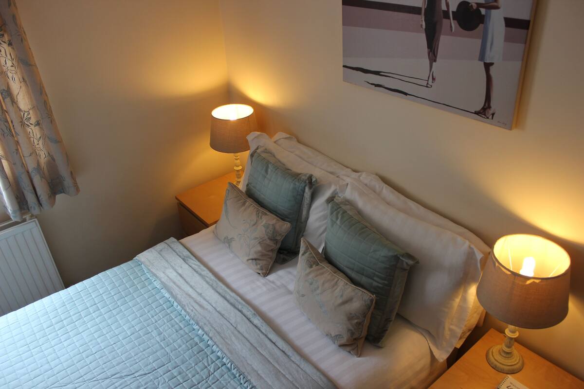 A neatly made bed with a blue quilt, two pillows, and two bedside lamps.