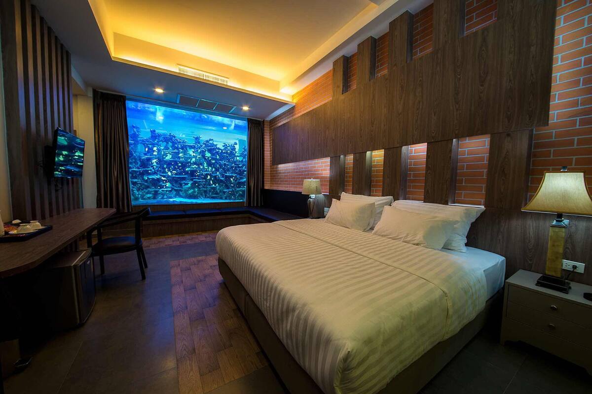 A hotel room with a large bed, a desk, a TV, and a view of a cityscape through a large window.