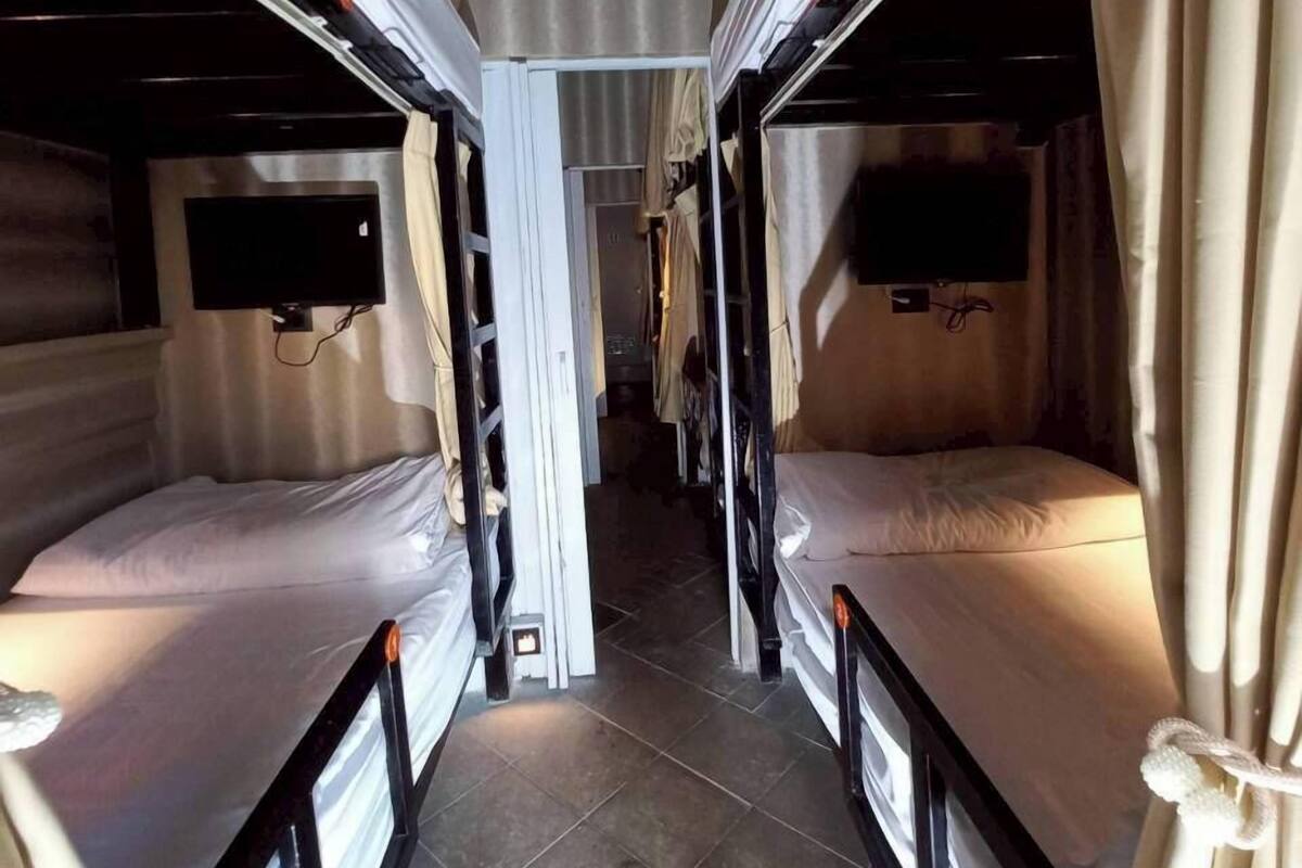 A narrow corridor with bunk beds on either side, a flat-screen TV mounted on the wall, and a tiled floor.
