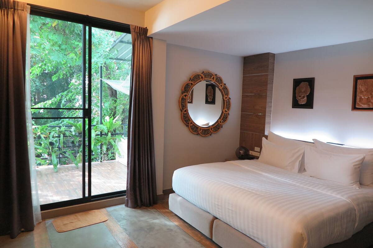 A bedroom with a large bed, a sliding glass door leading to a garden, a wooden headboard, and a mirror on the wall.