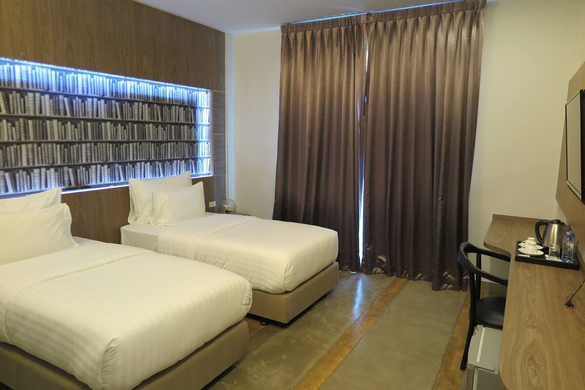 A hotel room with two beds, a desk, a chair, a bookshelf, and a large window with curtains.