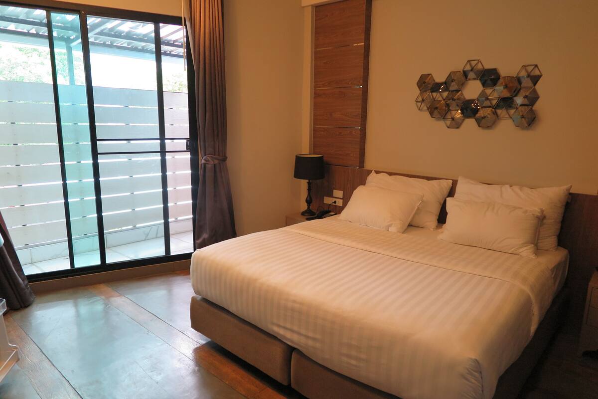 A bedroom with a large bed, a nightstand with a lamp, a wooden headboard, and a decorative wall piece.