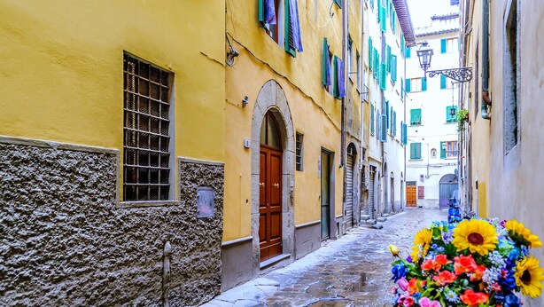 Una estrecha calle adoquinada con edificios amarillos, una puerta de madera y un gran arreglo floral.
