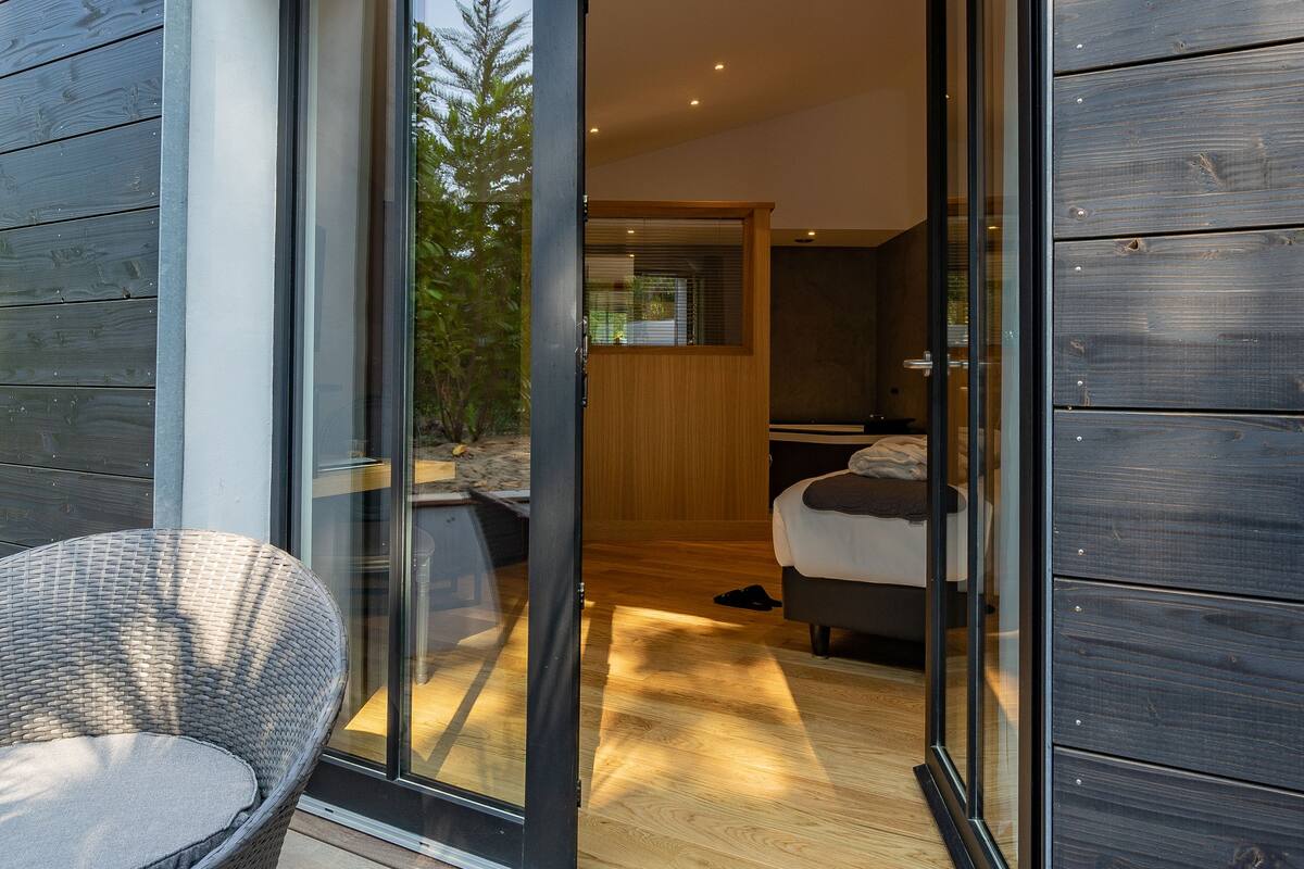 Un interior moderno con un vidrio que da a un dormitorio, una silla de mimbre afuera y una terraza de madera.