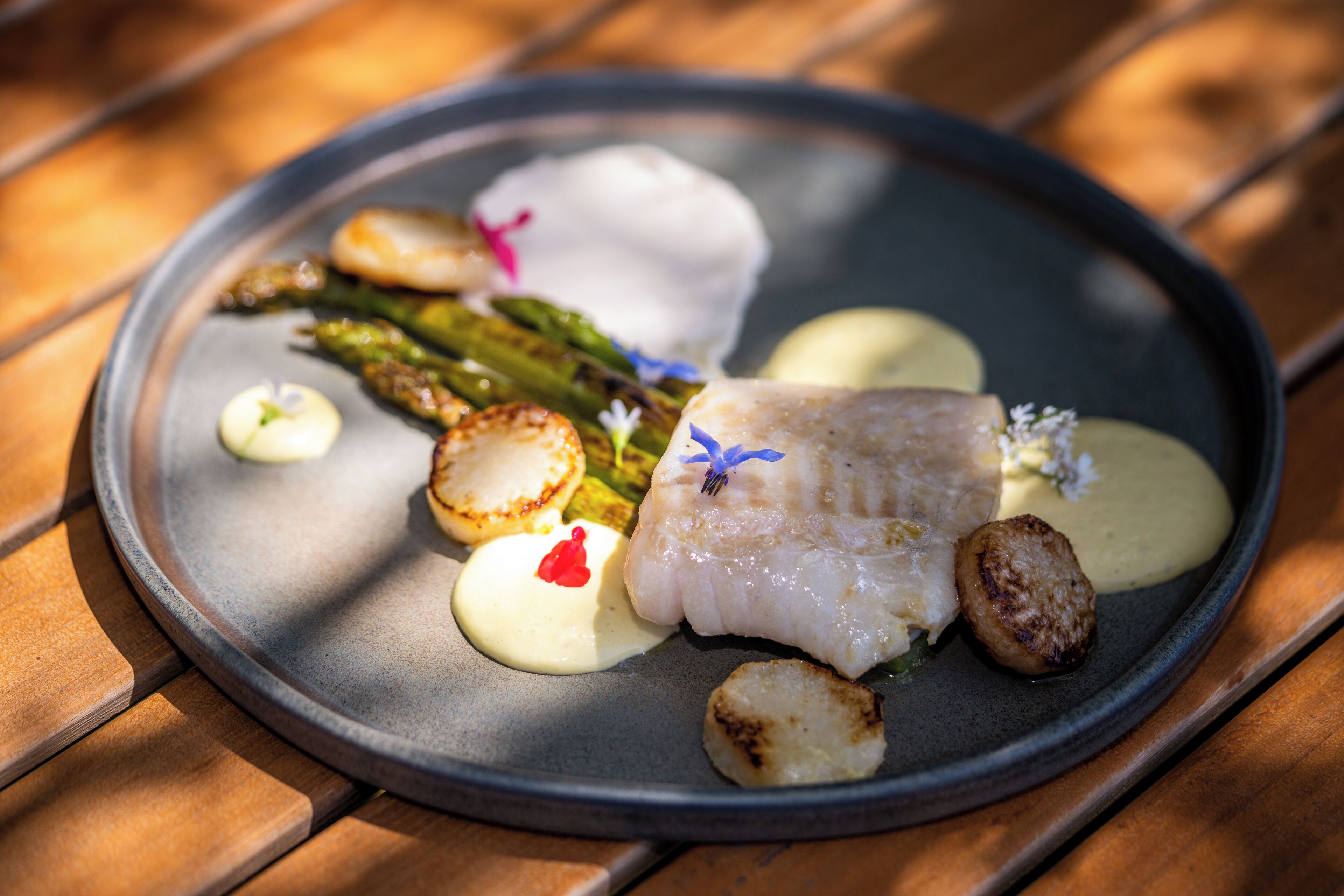 A plate of food with a piece of white fish, asparagus, and small round items, possibly scallops, on a dark plate.