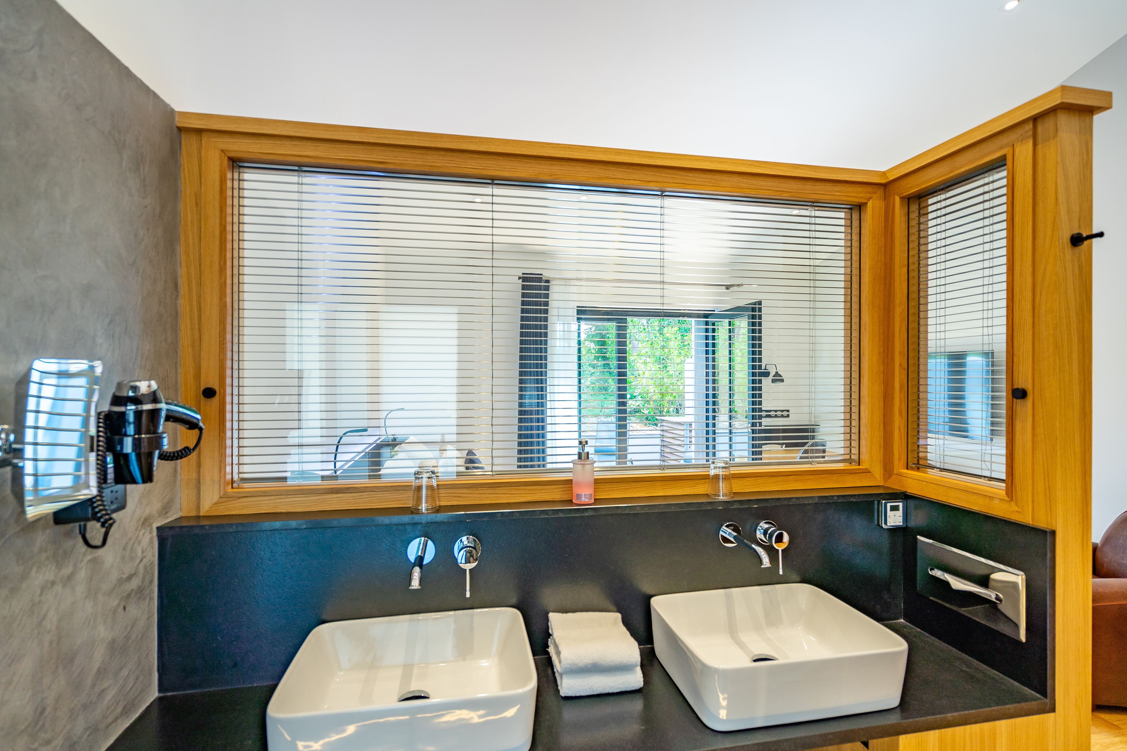 A modern bathroom with two sinks, a large mirror, and a window with blinds.