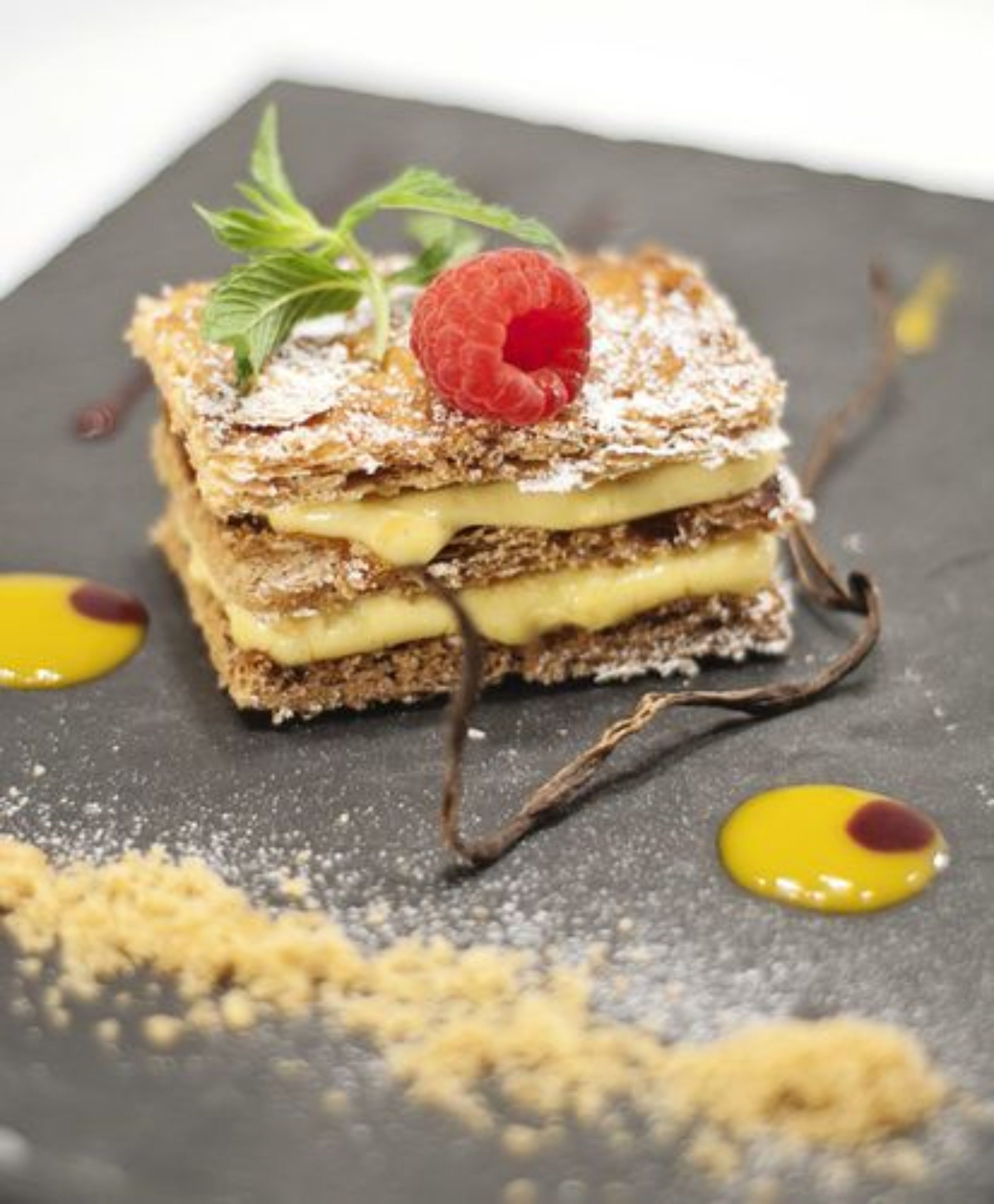 Un postre en capas con frambuesa encima, espolvoreado con azúcar glas y acompañado de rizos de chocolate y salsa amarilla.