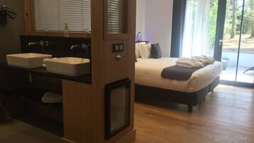 Una habitación de hotel moderna con suelo de madera, una cama grande con cabecera oscura, una mesita de noche blanca y un lavabo empotrado con espejo.