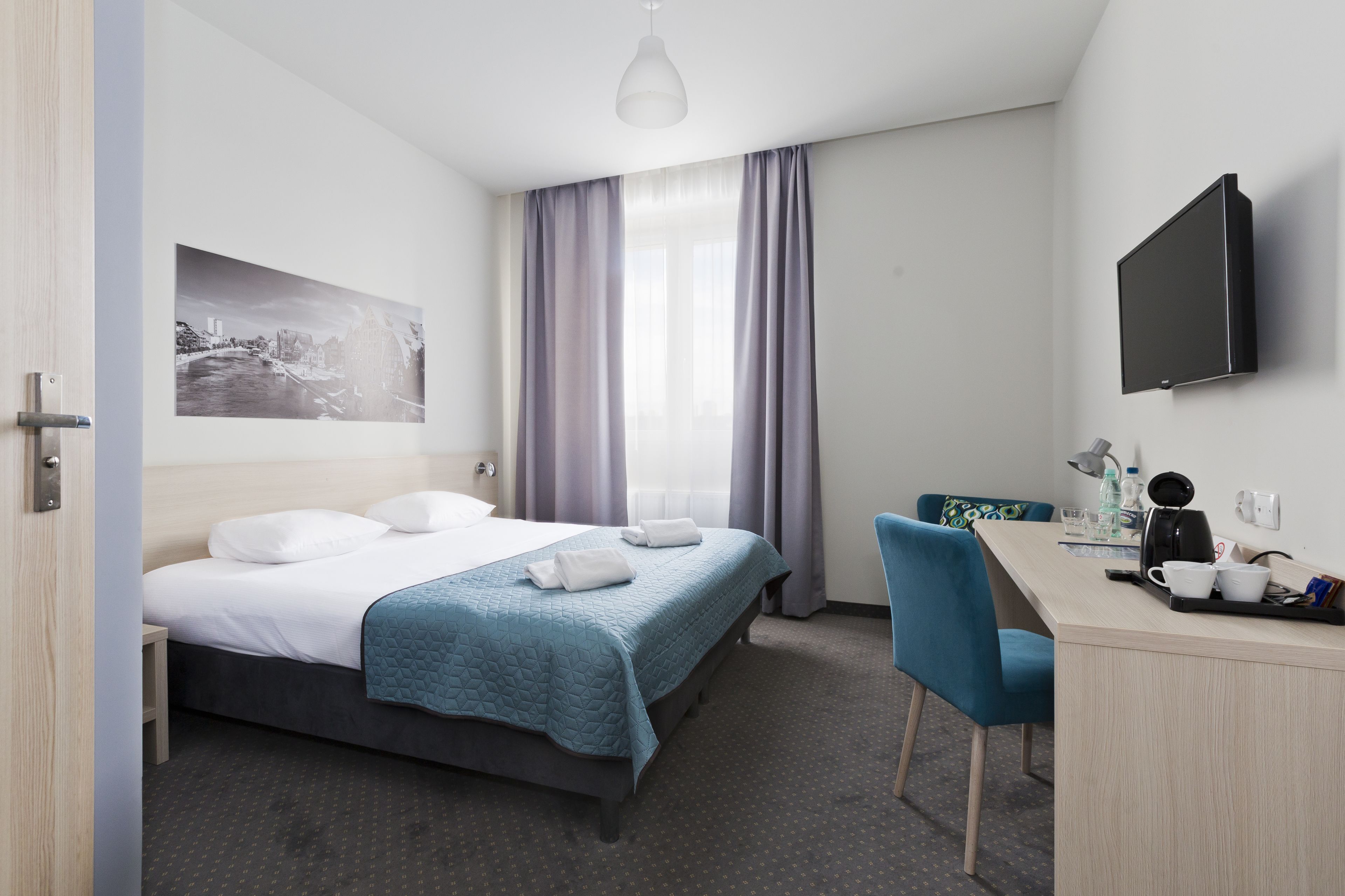 A hotel room with a bed, a desk, a chair, a flat-screen TV, a window with curtains, and a painting on the wall.