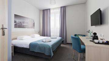 A hotel room with a bed, a desk, a chair, a flat-screen TV, a window with curtains, and a painting on the wall.