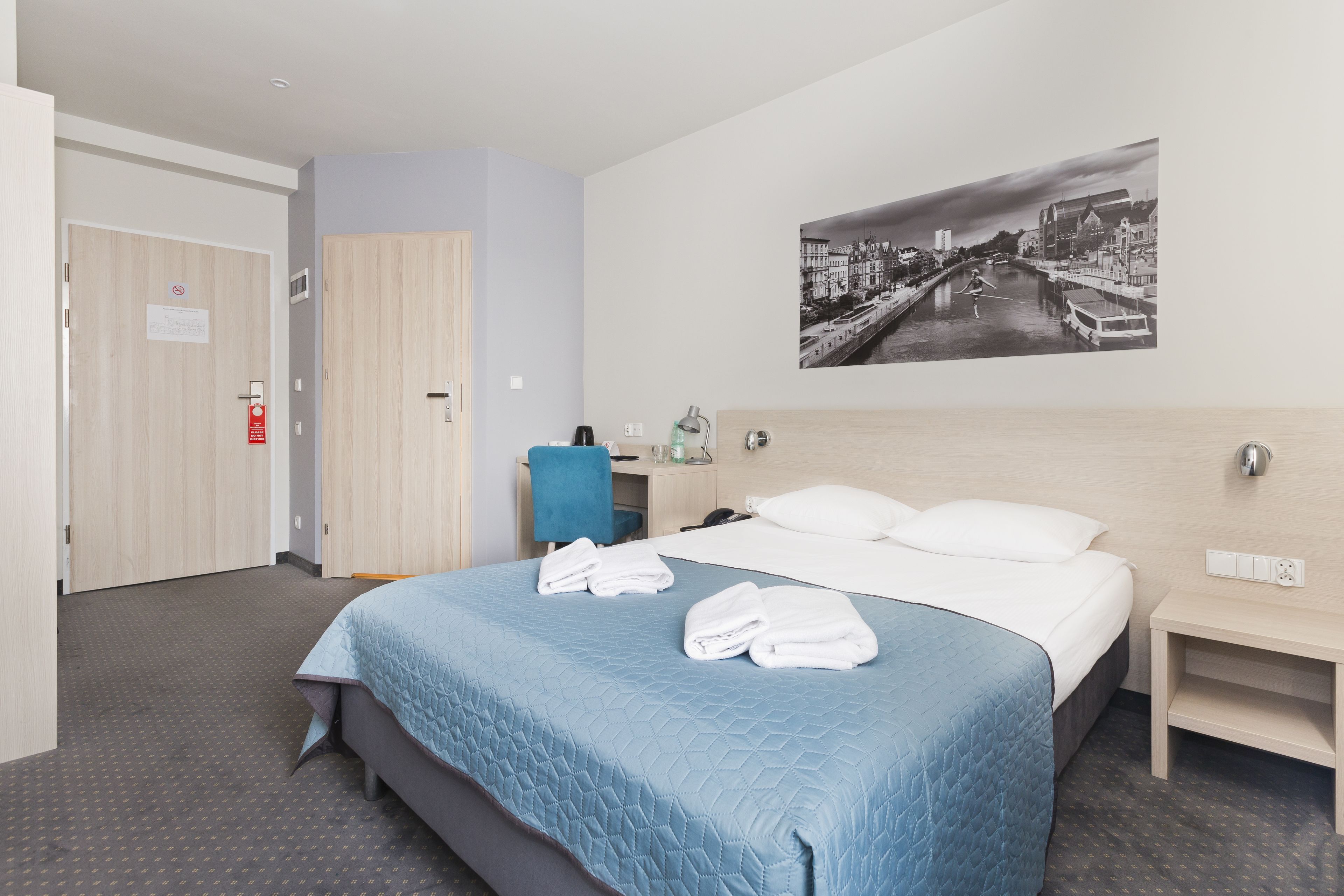 Une chambre d’hôtel comprenant un lit, un bureau, une chaise, une table de chevet et un tableau accroché au mur.