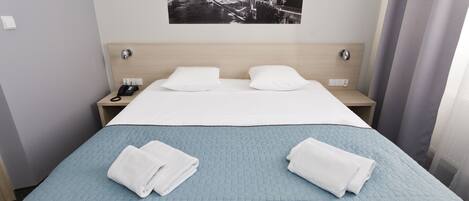 A hotel room with a large bed, two pillows, a nightstand with a phone, and a cityscape picture on the wall.