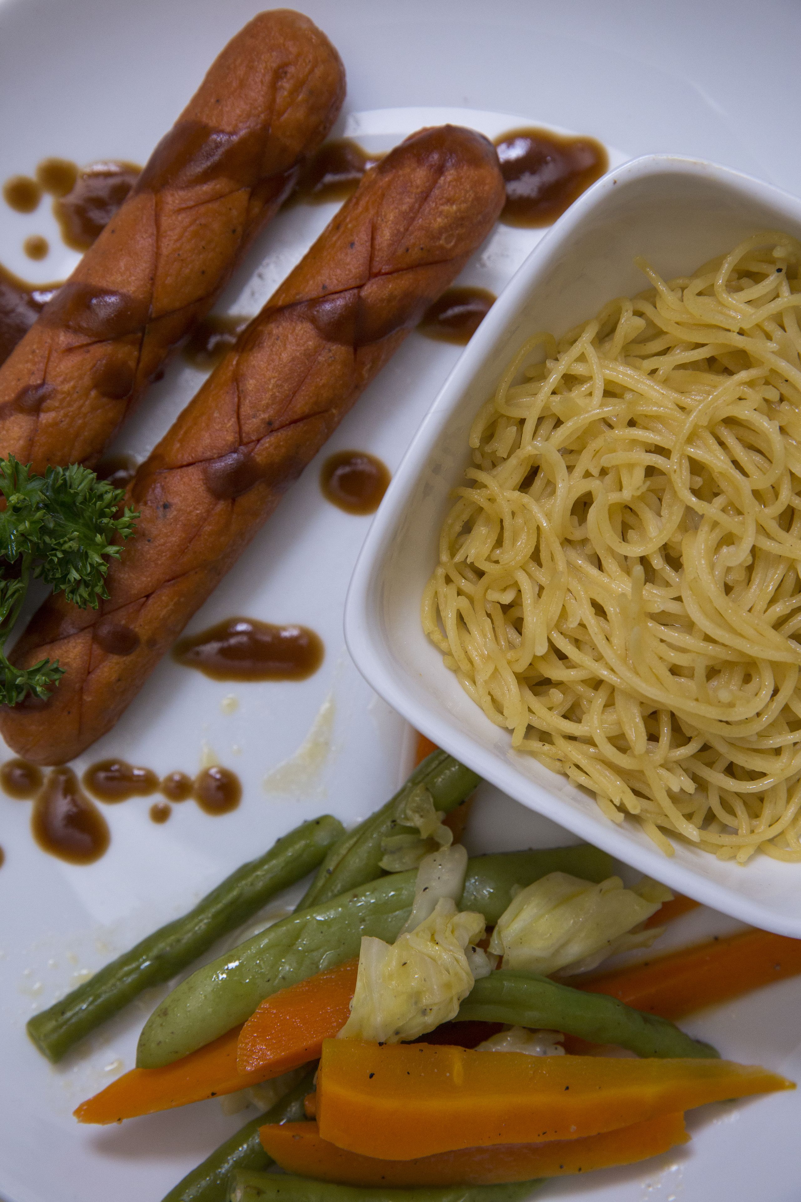 Um prato com duas salsichas grelhadas, uma porção de espaguete e uma mistura de legumes, incluindo vagem, cenoura e repolho.