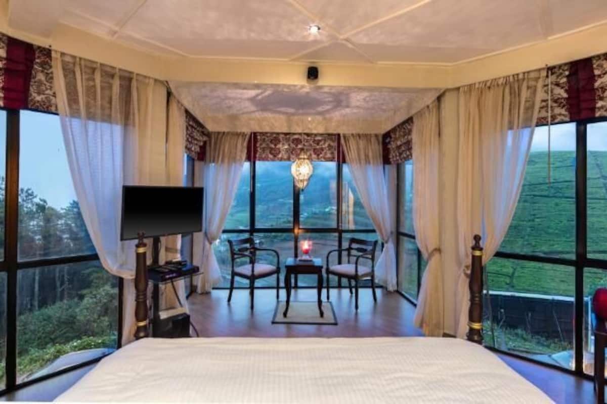 Une chambre avec un grand lit, une télévision, un bureau et une chaise. De grandes fenêtres offrent une vue sur la campagne.