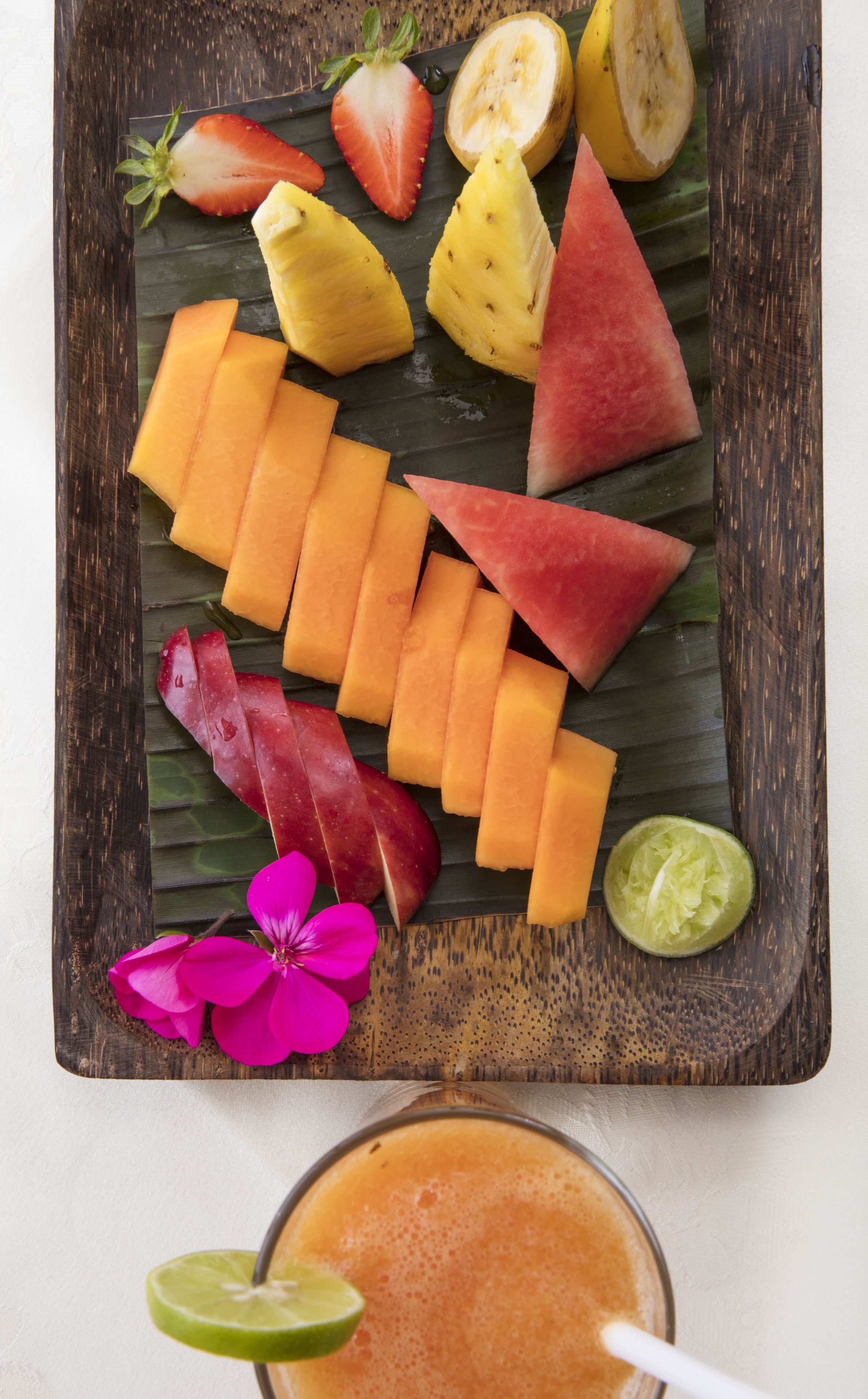 A wooden tray with various fruits and a flower on a white surface.