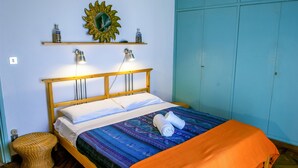 Un lit bien fait, recouvert d’une couvre-lit à motifs, deux lampes de chevet, un miroir en forme de soleil et une petite étagère avec des bouteilles.