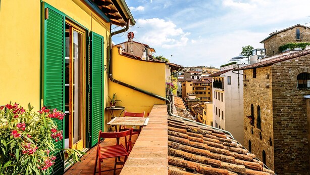 Une rue étroite, avec un immeuble jaune sur le côté gauche, une table et des chaises rouges sur un petit patio, et un escalier raide qui descend vers la rue.