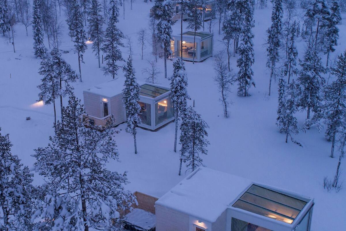 Moderni, lasikatolla varustettu mökki, jonka ympärillä on lumipeitteisiä puita.