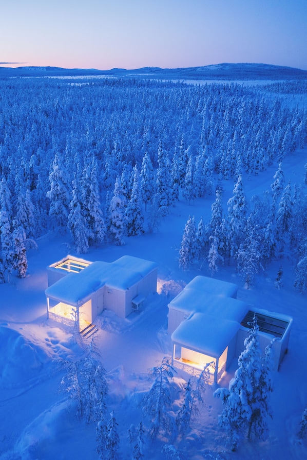 A snow-covered landscape with two cabins and a forest.