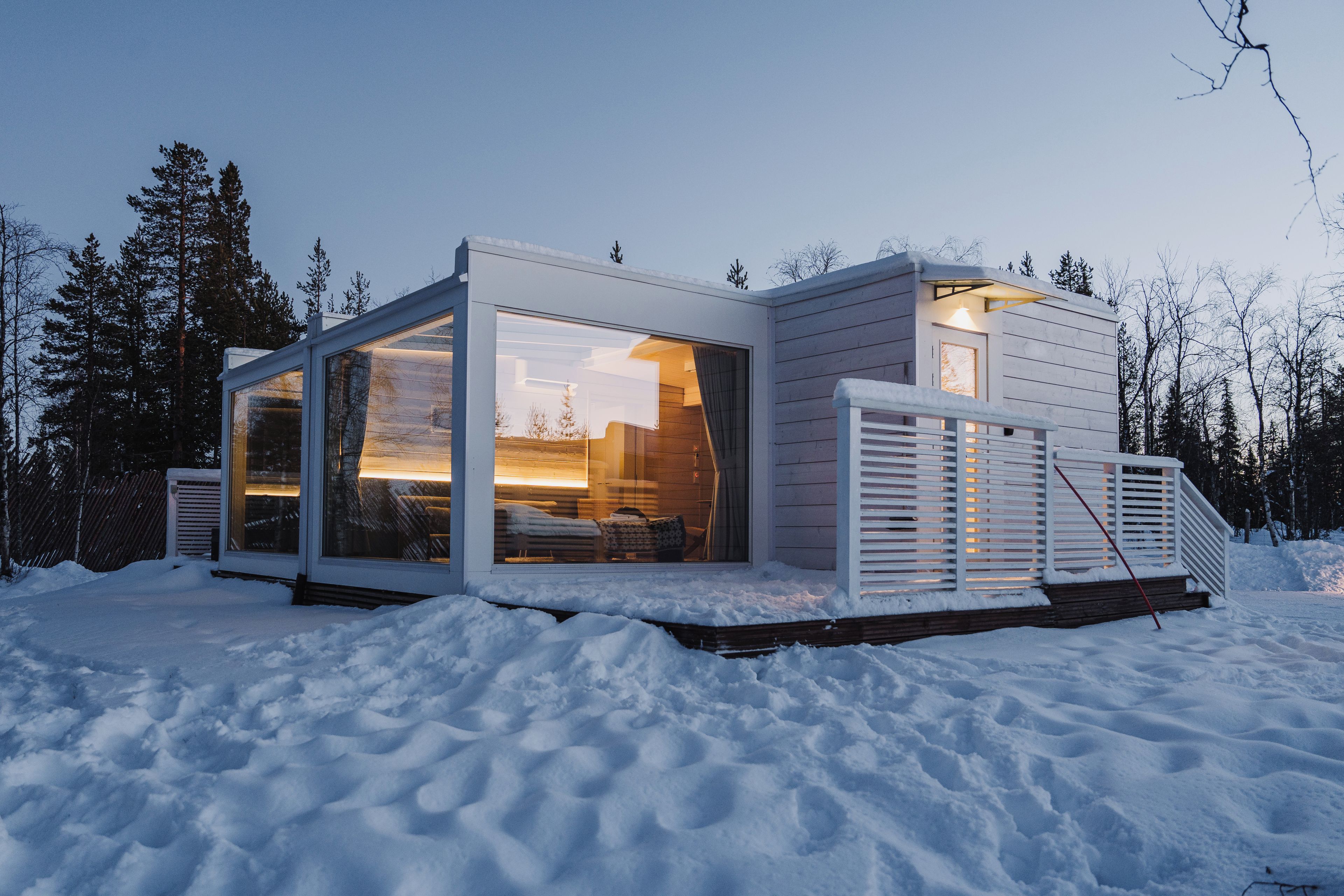 En moderne hytte med store glasruder, en træterrasse og et sneklædt landskab.