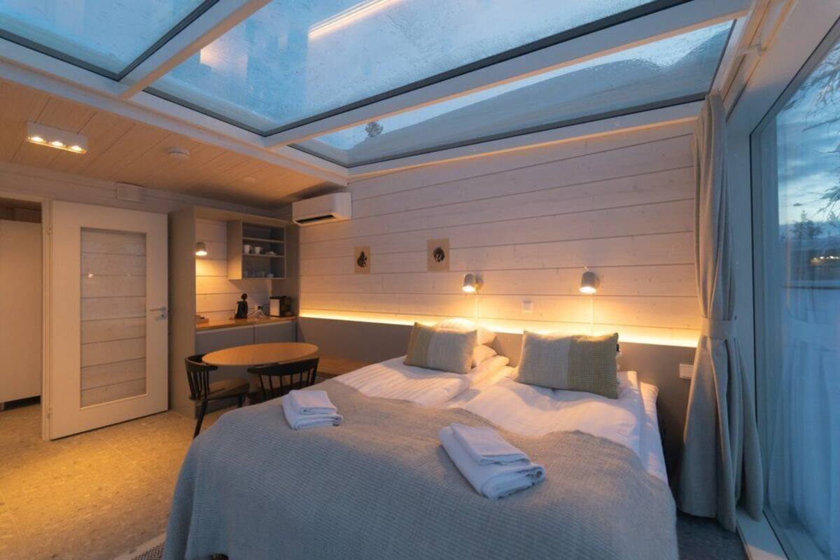 Moderni hotellihuone, jossa on kattovalokuvio, siististi taitelluilla pyyhkeillä varustettu sänky, pieni ruokapöytä ja sisäänrakennettu keittiöalue.
