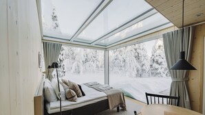 Ein modernes Interieur mit Glasdecke, einem Bett mit Kissen, einem Holztisch und Blick auf eine verschneite Landschaft.