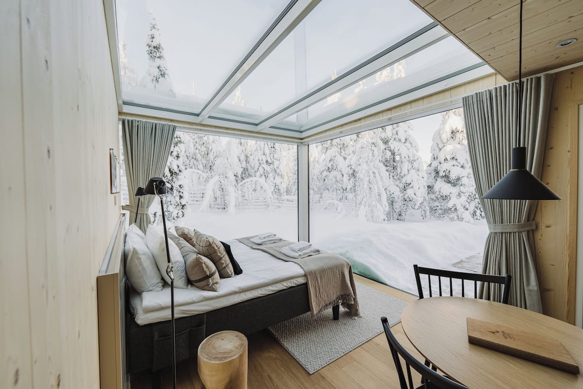 Moderni sisustus, jossa on lasikatto, tyynyillä varustettu sänky, puinen pöytä ja näkymä lumiseen maisemaan.