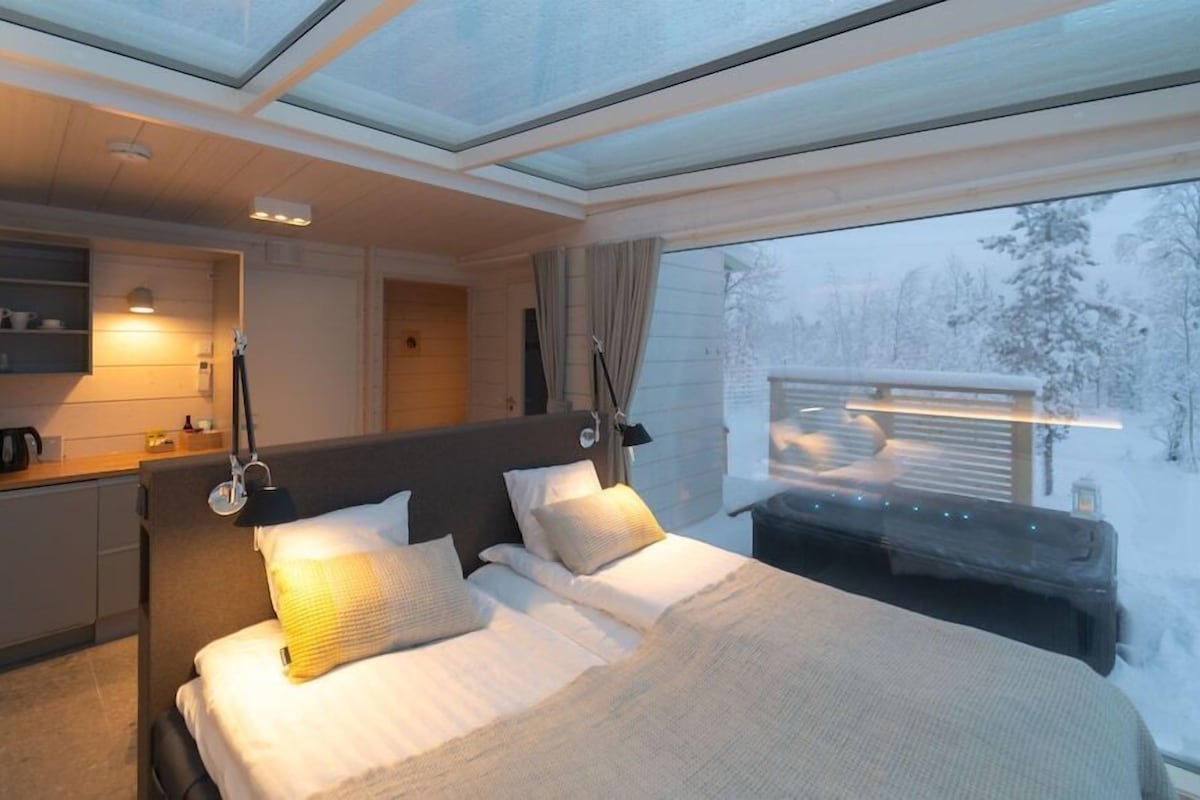 A modern hotel room with a large bed, bedside lamps, a small desk, and a view of a snowy forest.